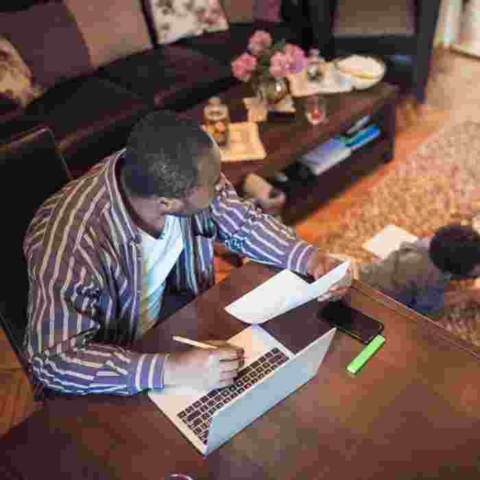 Man sat at desk at home with a laptop and a piece of paper while two children play on the rug behind him