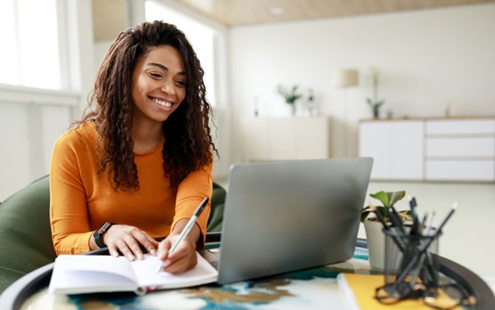 Person sat at table smiling at a laptop and taking notes